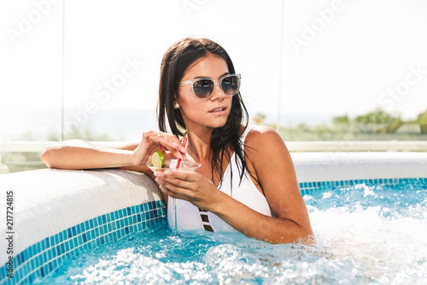 Fototapeta Portrait of sexual european woman in white bathing suit and sunglasses sitting in swimming pool, and drinking cocktail outdoor in spa resort