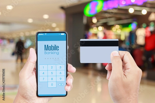 Fototapeta Hand holding smartphone with Mobile Banking on screen over blurred in shopping mall background for online payment via credit card