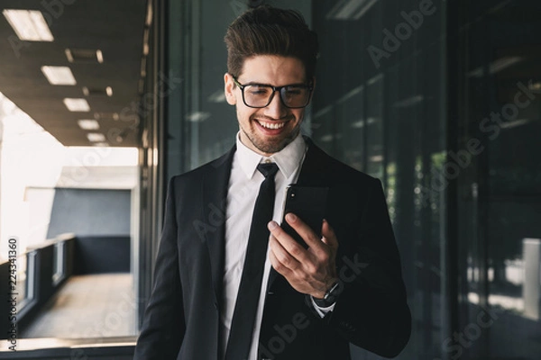 Fototapeta Portrait of handsome young businessman dressed in formal suit standing outside glass building, and holding mobile phone