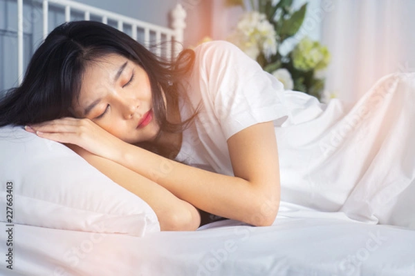 Fototapeta Woman stretching in bed after waking up, back view. Woman sitting near the big white window while stretching on bed after waking up with sunrise at morning, back view.