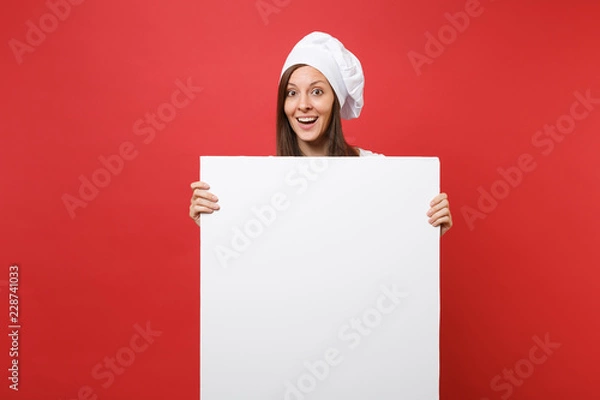 Fototapeta Housewife female chef cook baker in striped apron, white t-shirt, toque chefs hat isolated on red background. Woman hold big white blank billboard for promotional content. Mock up copy space concept.