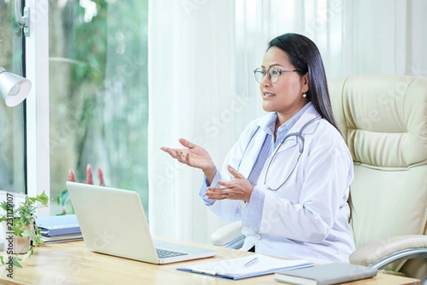 Fototapeta Adult Thai woman in white coat gesturing while speaking at table in hospital office having consultation