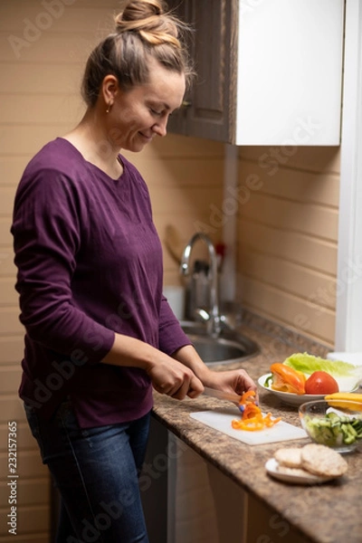 Fototapeta A young woman cuts vegetables and prepares a salad in the kitchen.