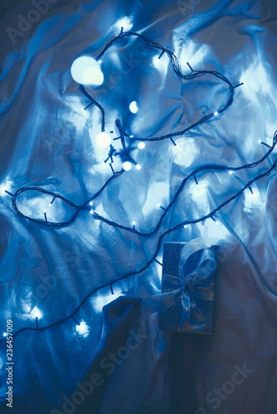 Fototapeta cropped shot of woman and wrapped christmas gift on bed with blue festive lights