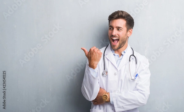 Fototapeta Handsome young doctor man over grey grunge wall smiling with happy face looking and pointing to the side with thumb up.