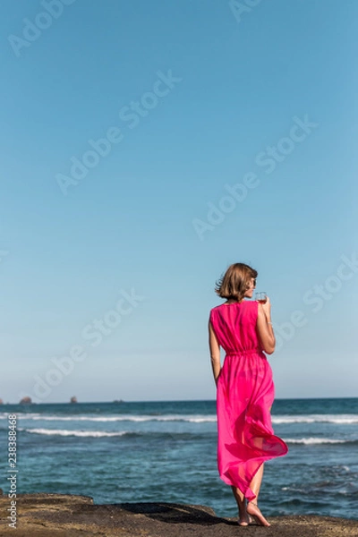 Fototapeta Young woman in a long red dress and with a glass of wine posing on the sea background.