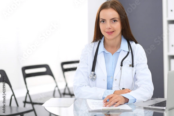 Fototapeta Doctor woman at work. Portrait of female physician filling up medical form while sitting at the glass desk at clinic or hospital. Medicine and healthcare concept