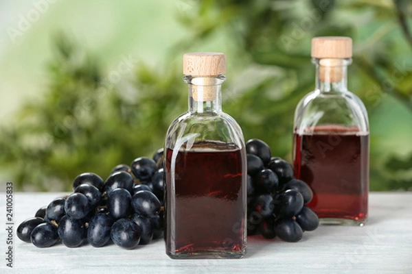 Fototapeta Bottles with wine vinegar and fresh grapes on wooden table against blurred background