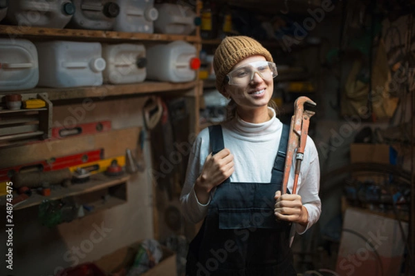 Fototapeta Portrait of smiling young builder girl standing in old workshop room, wearing construction glasses.