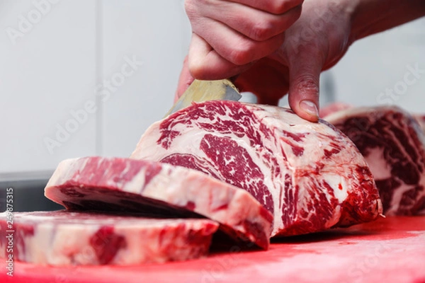 Fototapeta Closeup macro cook's hand cut ribeye marbled beef steak with knife on red plastic cutting board on metal table in restaurant kitchen. Concept packing semifinished products, steakhouse, grill meat