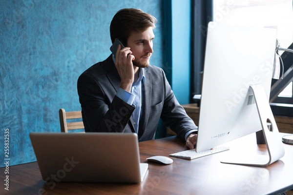 Fototapeta Portrait of handsome young male sitting at office desk with laptop computer and talking on mobile phone. Communication concept.