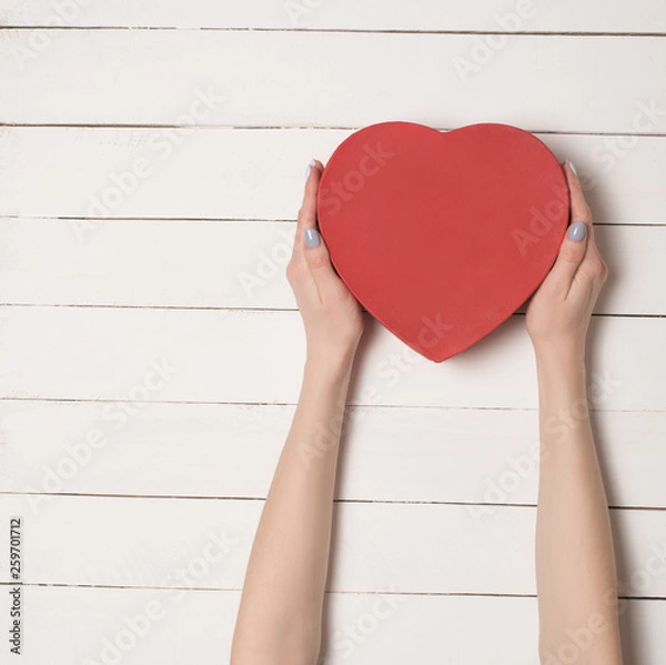 Fototapeta Female hands hold a red heart shaped box against the background of a white wooden table