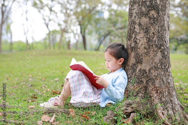 Fototapeta Pretty little girl reading a book sitting under a tree outdoor garden at summer day.