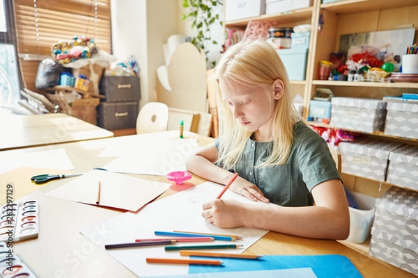 Fototapeta Adorable albino girl drawing on white paper sheet with colored pencils while sitting at table in art school