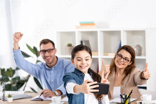 Fototapeta selective focus of cheerful kid taking selfie on smartphone near happy parents gesturing in office