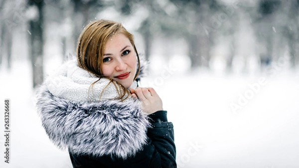 Fototapeta Portrait of a beautiful girl in the black jacket with fur hood amid winter forest