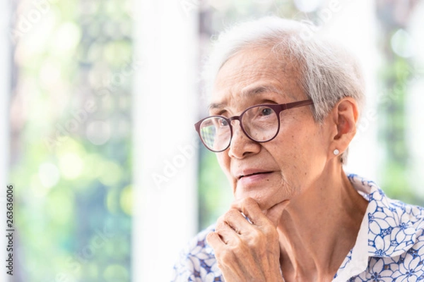 Fototapeta Asian elderly woman in glasses,thinking with hand on chin in her home,senior woman smiling feeling happy