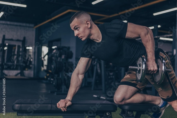 Fototapeta Handsome muscular man working out with dumbbells at the gym. Attractive athletic man with strong ripped body lifting weights at sport studio, copy space. Motivation, confidence concept