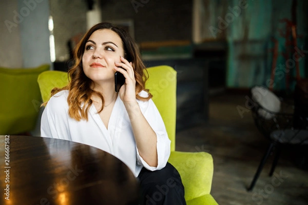 Fototapeta Beautiful smiling young woman dressed in dress sitting in chair at the cafe, talking on mobile phone, stylish interior