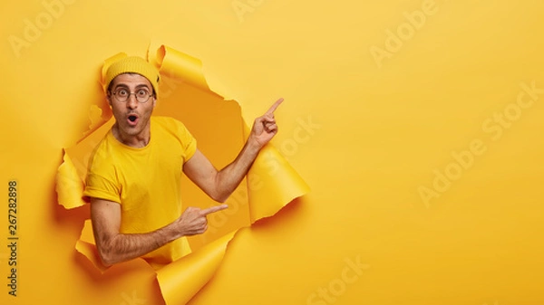 Obraz Indoor shot of impressed delighted unshaven man points away at upper right corner, poses through torn paper hole, wears yellow hat and t shirt, advertises awesome product. Advertisement concept