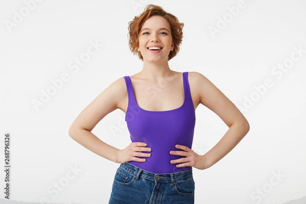 Fototapeta Portrait of a happy young attractive short-haired girl, wearing a purple jersey and jeans, holding hands on her waist, smiling broadly and looking at the camera isolated over white background.