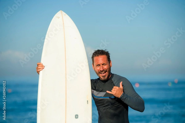 Fototapeta handsome and attractive surfer man in neoprene swimsuit holding surf board posing cool after surfing enjoying Summer water sport and holidays giving hand sign