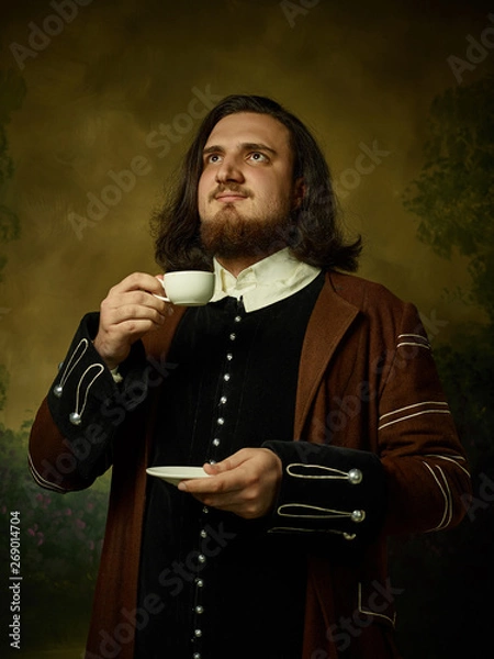 Fototapeta Young man as a medieval knight on dark studio background. Portrait in low key of male model in retro costume. Drinking morning coffee. Human emotions, comparison of eras and facial expressions concept