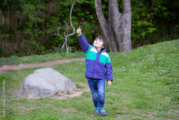 Fototapeta Active boy pointing wooden stick up  while walking in forest park,Child boy having fun playing outdoor activity,Preschool Kid wearing acket adventure in spring or summer forest on school trip