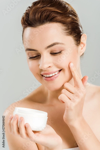 Fototapeta happy woman looking at container while applying face cream isolated on grey