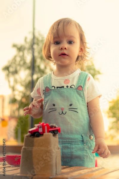 Fototapeta Little smiling girl playing with sand castle and red car toy in sandbox