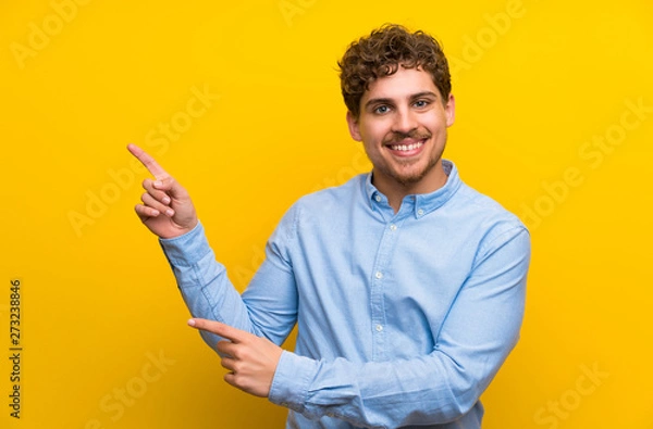 Fototapeta Blonde man over isolated yellow wall pointing finger to the side