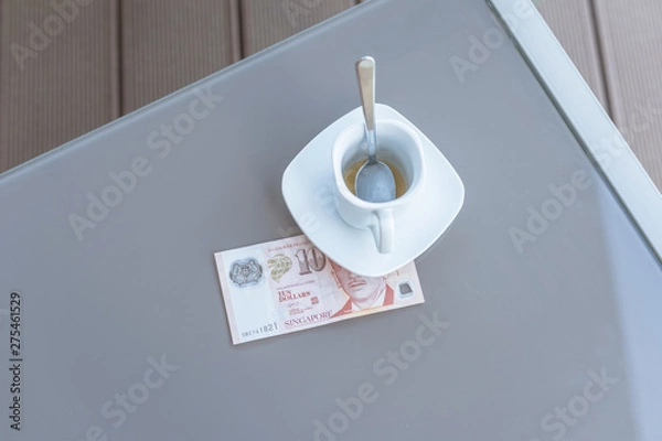 Fototapeta Singapore ten dollars bill and empty cup of coffee on a glass table of outdoor cafe. Payment, tip.