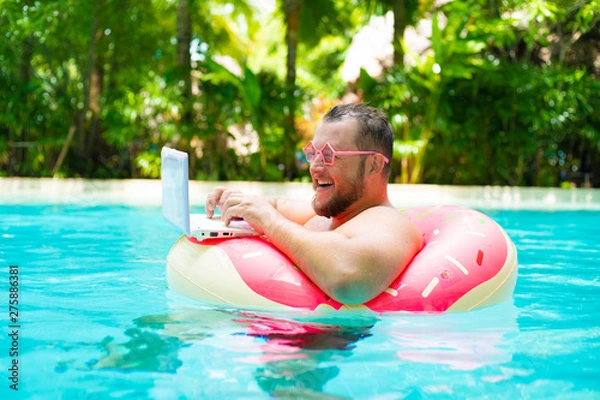 Fototapeta Funny fat male in pink glasses on an inflatable circle in the pool works on a laptop portraying a girl.