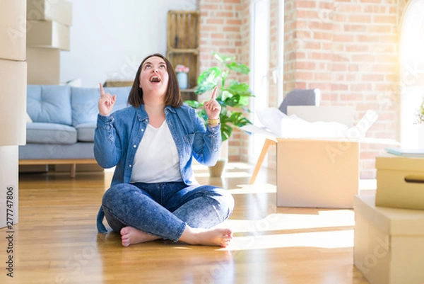 Fototapeta Young plus size woman sitting on the floor around cardboard boxes moving to a new home amazed and surprised looking up and pointing with fingers and raised arms.