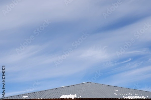 Fototapeta The roof of the metal profile with melting snow against a blue sky with clouds