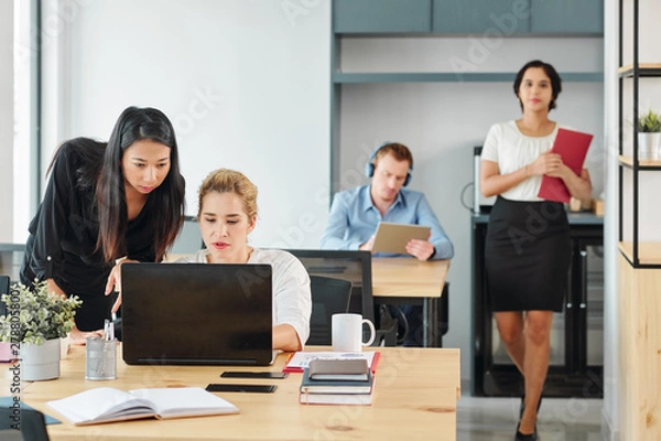 Fototapeta Group of young business people sitting at their workplaces and working with laptop computers with young businesswoman walking along the office corridor