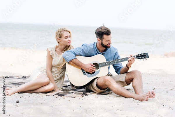 Fototapeta beautiful young barefoot woman sitting on blanket with boyfriend playing acoustic guitar at beach near sea