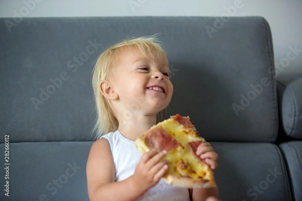 Fototapeta Cute little Caucasian kid eating pizza. Hungry child taking a bite from pizza on a pizza party, outdoors