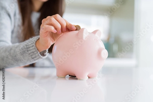 Fototapeta Young woman putting a coin inside piggy bank as savings for investment