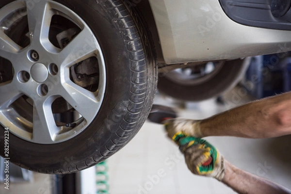 Fototapeta Car repair in the service station. Hands of a mechanic in overalls repairing the car on the lift without wheel, holding the tire and mechanical works.