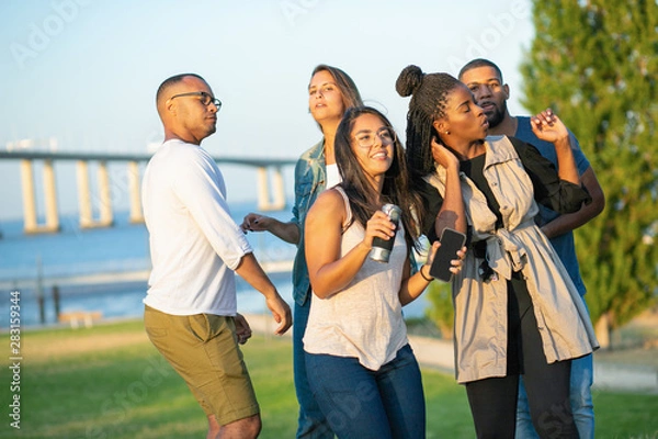 Fototapeta Company of young people dancing at park. Friends listening music from smartphone. Relaxed friends spending time together. Concept of party