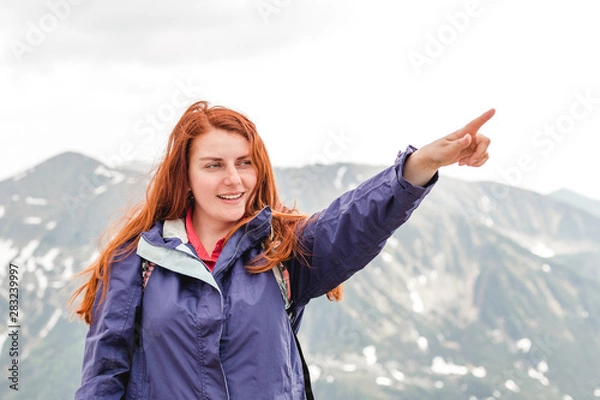 Fototapeta Young woman with backpack pointing at something in the distance. Trekking and tourism concept. Copyspace. A young pretty redheaded woman standing on a background of mountains