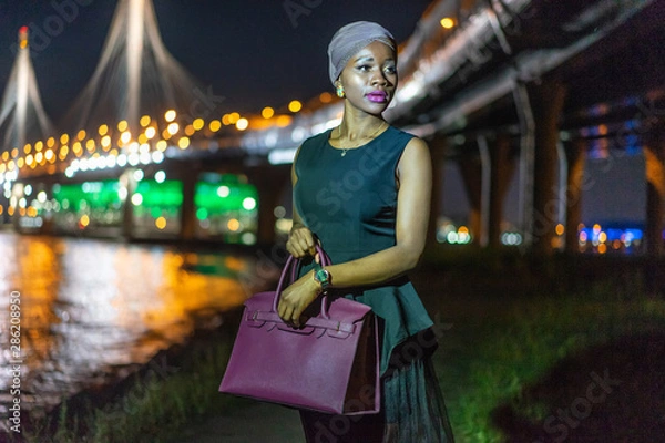 Fototapeta A dark-skinned girl in a black dress, a purple turban and a purple bag. Model posing against the background of the evening city. The woman returns home late. Night life. Lights of the night city.