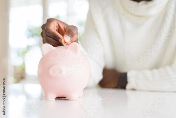 Fototapeta Close up of african man hands putting a coin inside piggy bank, saving money as investment