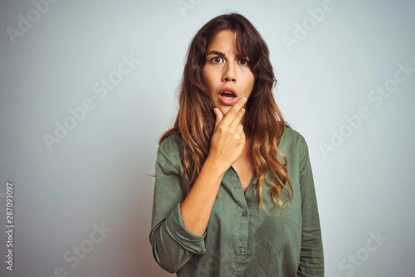 Fototapeta Young beautiful woman wearing green shirt standing over grey isolated background Looking fascinated with disbelief, surprise and amazed expression with hands on chin