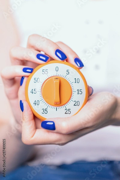 Fototapeta Female hands with blue manicure holding kitchen timer on a light background. Time control concept