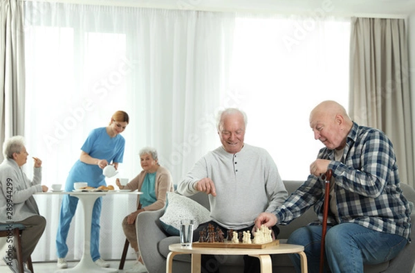 Fototapeta Elderly men playing chess while nurse serving breakfast to women at retirement home. Assisting senior people