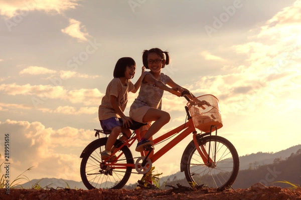 Fototapeta Silhouette two children playing riding bike on mountain at sunset sky background