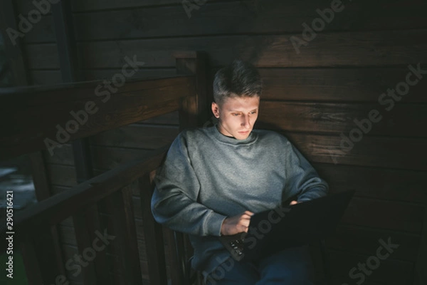 Fototapeta Portrait of handsome young man sitting on balcony in chair and using laptop, looking at screen with serious face. Guy works on a laptop in the evening in the apartment. Freelancer works at home.