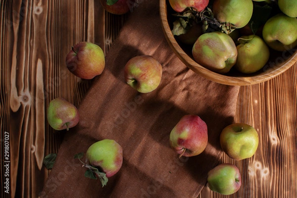 Fototapeta Top view of green and red apples on wooden table and brown linen cloth and wooden bowl full of fresh organic fruits. Autumn harvest set